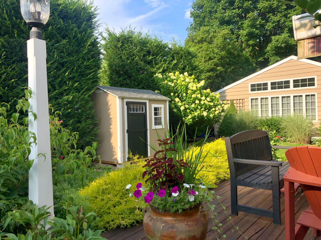 Garden scene with wooden shed, landscaped plants, bench, and colorful flowers on a sunny day.
