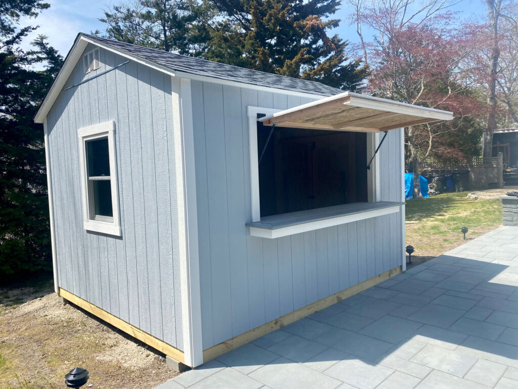 Gray outdoor shed with open serving window, situated on a patio, surrounded by trees and sunlight.