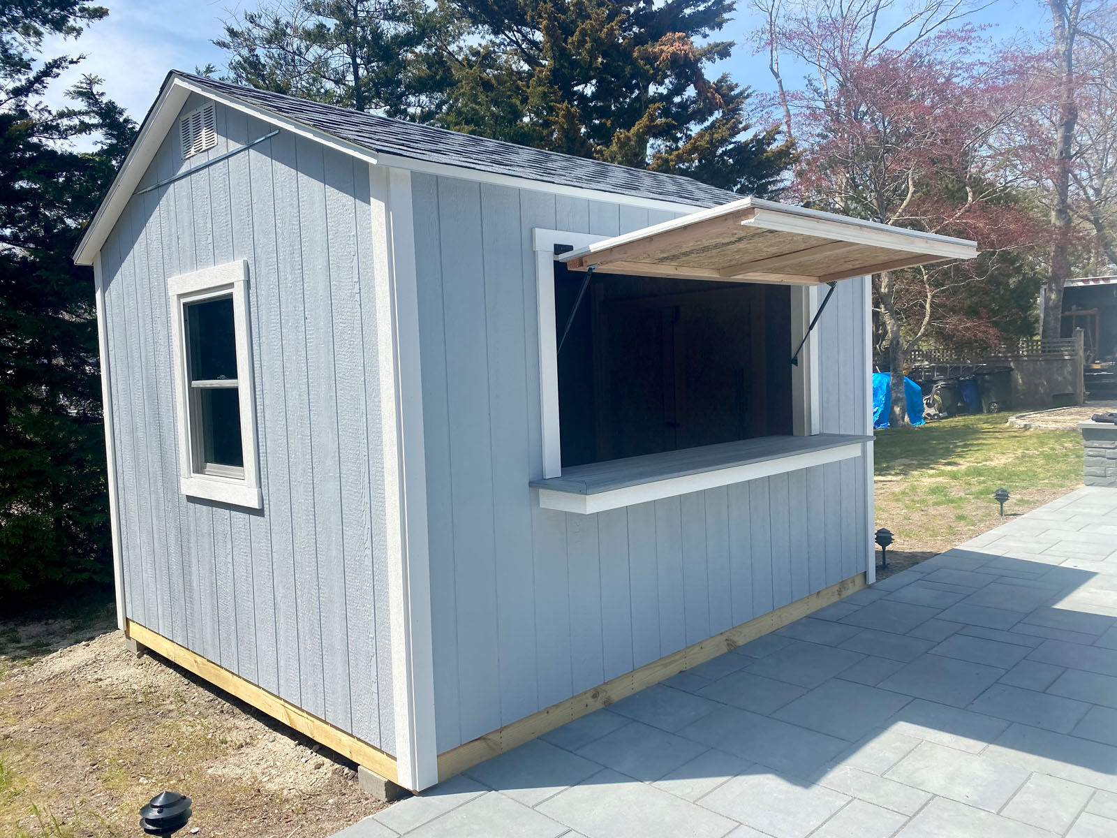 Gray outdoor shed with open serving window, situated on a patio, surrounded by trees and sunlight.
