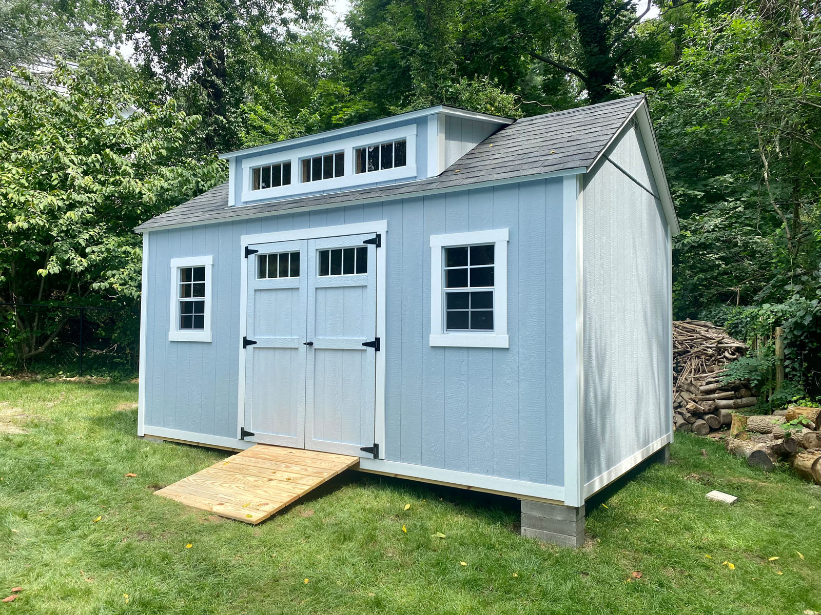 Light blue backyard shed with double doors, surrounded by lush greenery and a woodpile, on a grassy lawn.