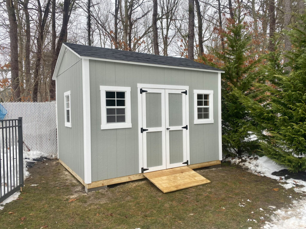 A gray garden shed with white trim and double doors, set in a snowy backyard with trees.