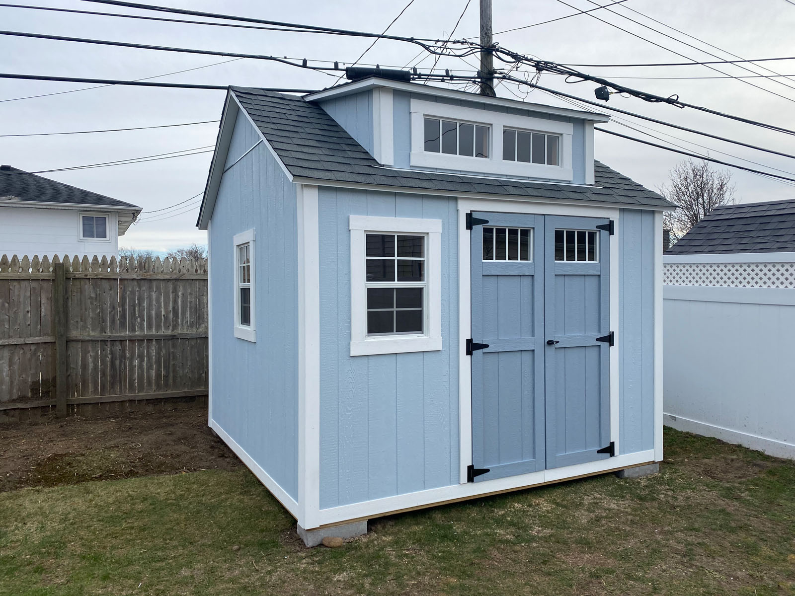 Light blue garden shed with double doors and windows, set on grass with a wooden fence background.