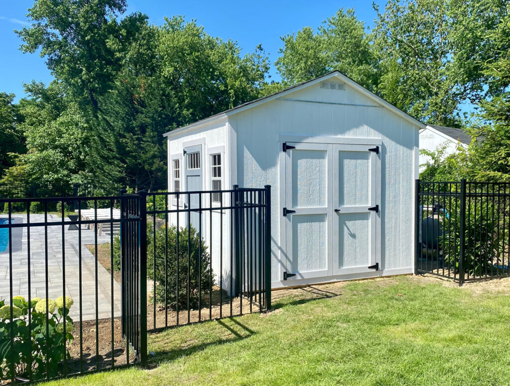 White garden shed with black hardware, surrounded by metal fence, lush greenery, and a glimpse of a tiled patio.