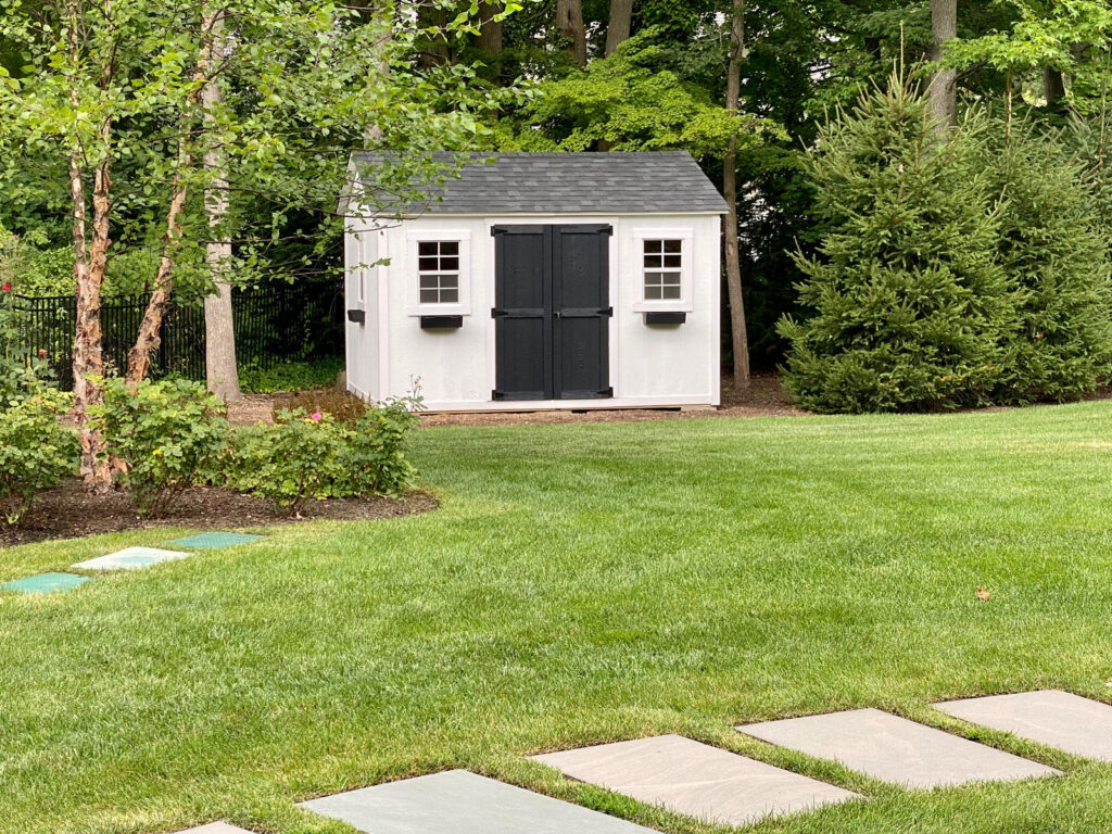 White garden shed with black doors, surrounded by green grass, trees, and shrubs in a landscaped backyard.