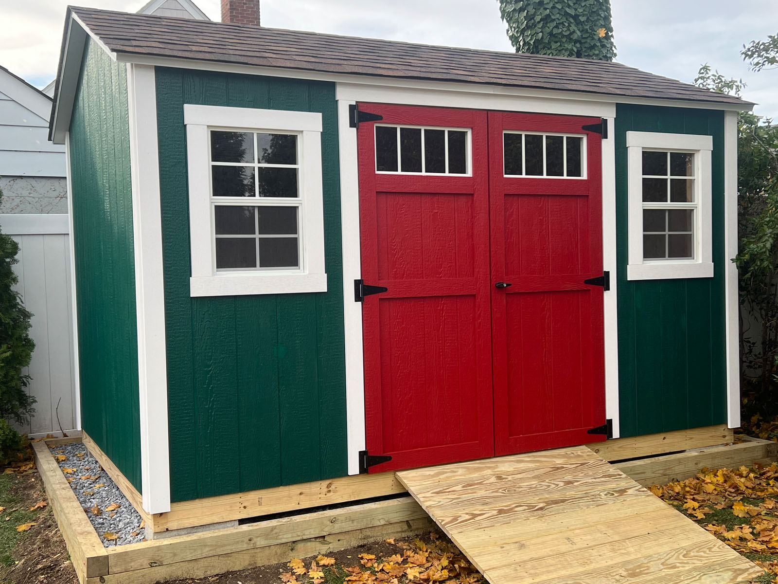 Green garden shed with red double doors and ramp, surrounded by autumn leaves.
