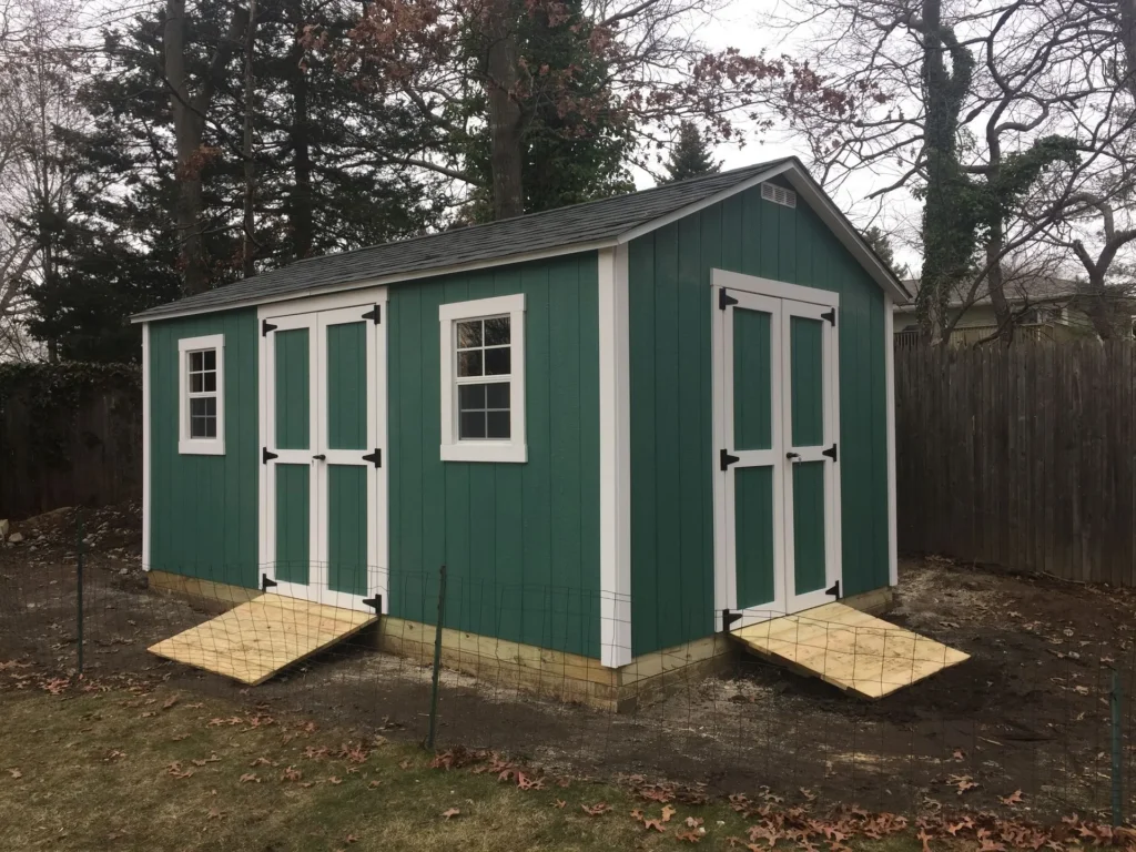 Green wooden garden shed with white trim, dual doors, and ramps in a fenced backyard surrounded by trees.