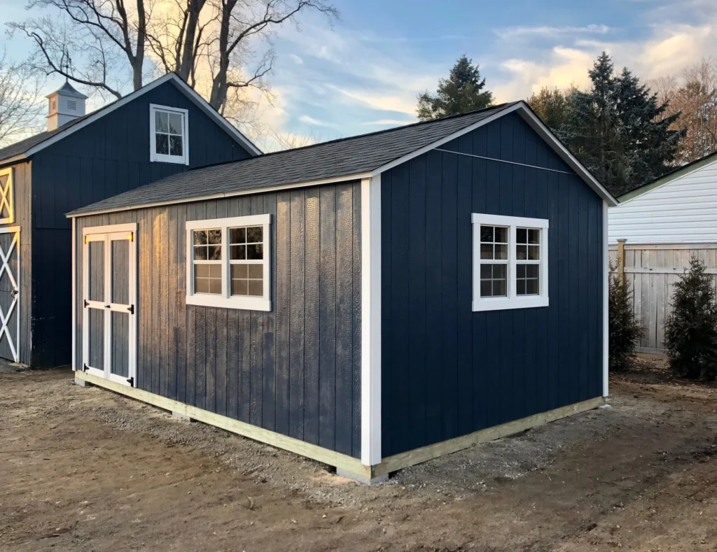 Blue wooden shed with white trim, double doors, and windows in a backyard setting under a clear sky.