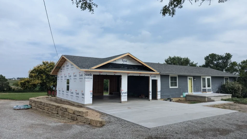 Newly constructed house with Tyvek wrap, concrete driveway, and a yellow door, set in a suburban landscape.