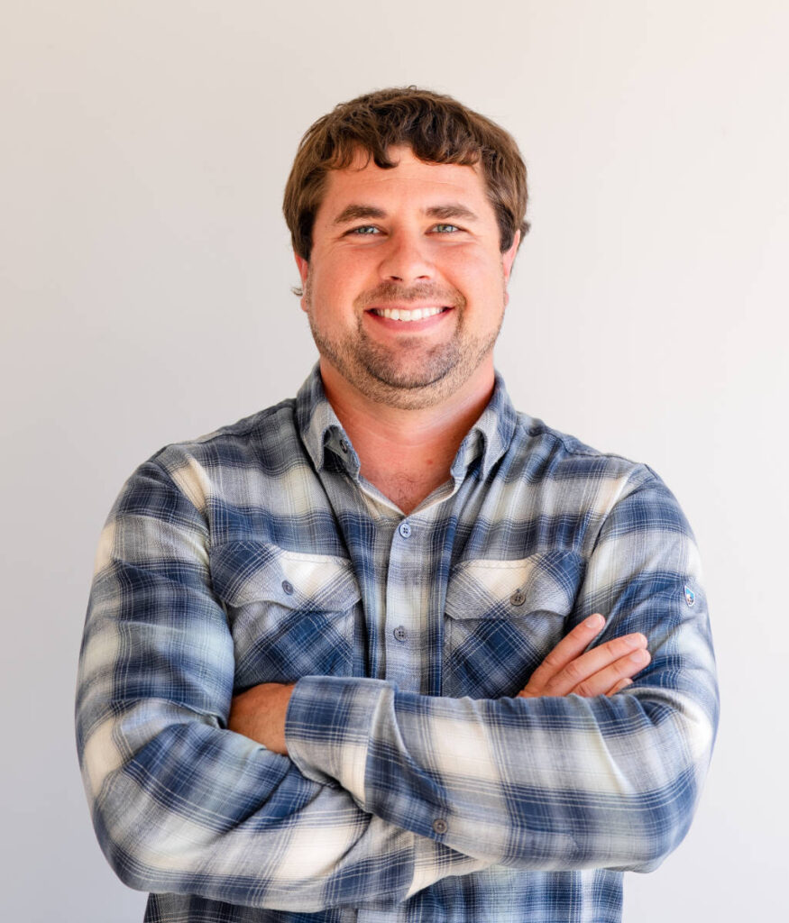 Smiling man in a blue plaid shirt with crossed arms against a light gray background.