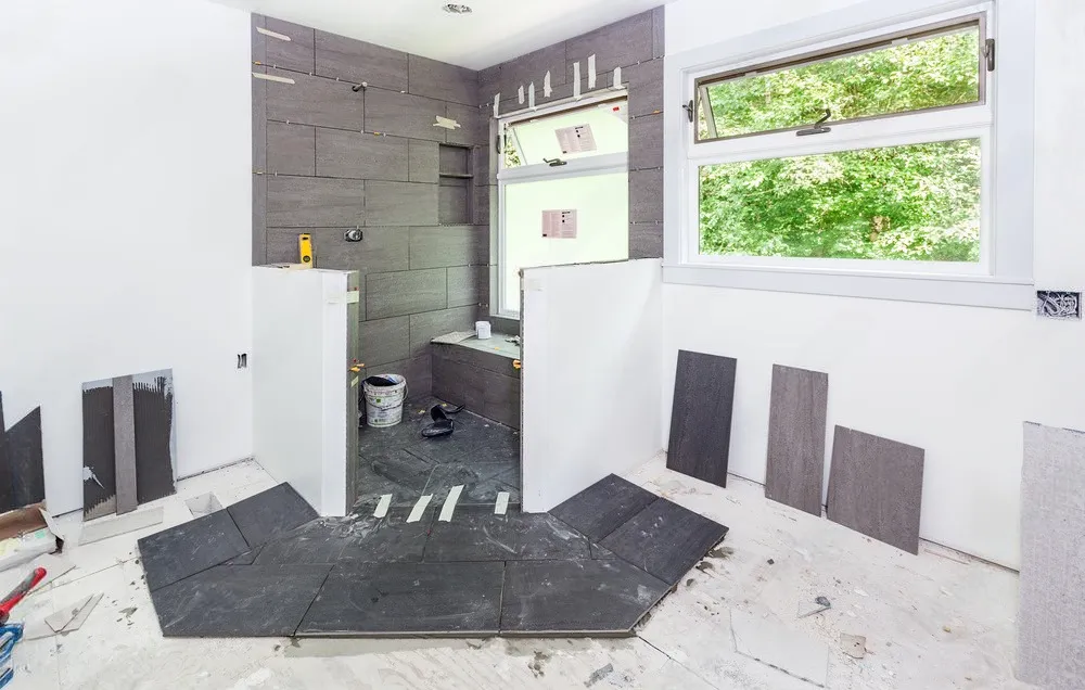 Bathroom under renovation with dark gray tiles, white walls, large window, and construction materials scattered throughout.