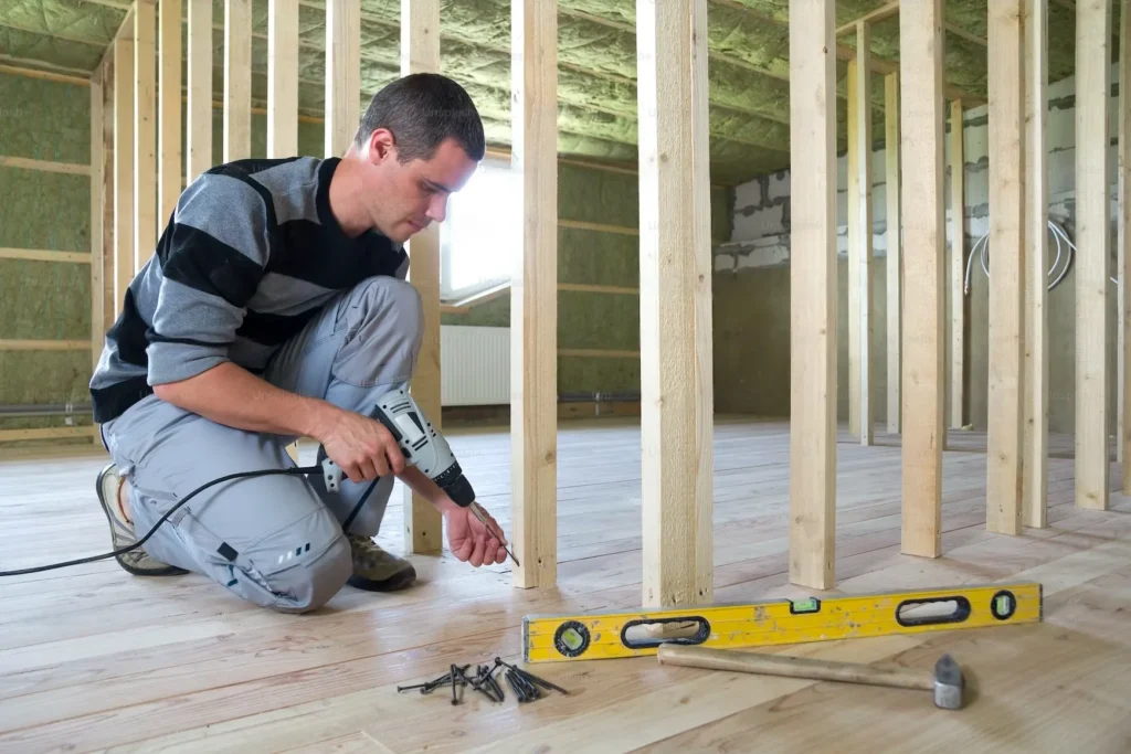 Man using a power drill on wooden framing in unfinished room, with level and hammer on floor.